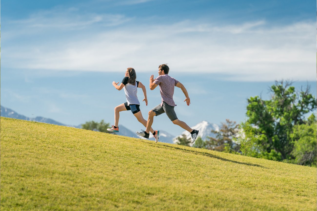 A man and woman in running attire, wearing specialized running shoes, sprinting up a steep hill with determination.